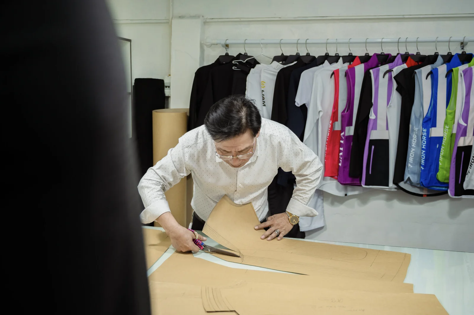 Iron Horse pattern maker cutting a paper garment pattern by hand in the Ho Chi Minh City sample room, with finished sportswear on the rail behind